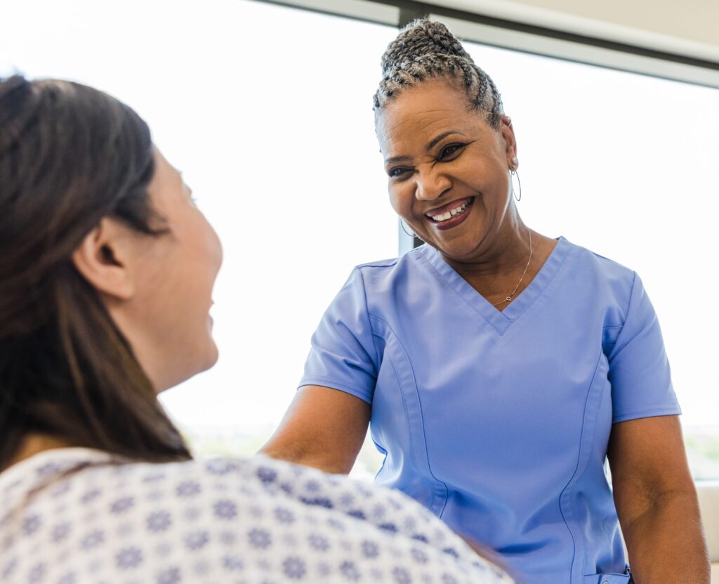 An unrecognizable female patient and a senior adult female nurse smile at each other.