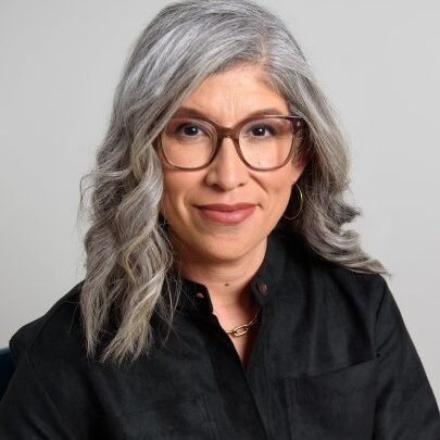 A woman with wavy gray hair, wearing large glasses, a black shirt, and gold hoop earrings, smiles softly while sitting against a plain light background.