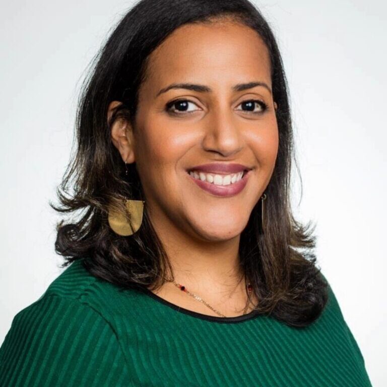 A woman with shoulder-length dark hair smiles at the camera. She is wearing a green top, gold earrings, and a necklace, with a light background.