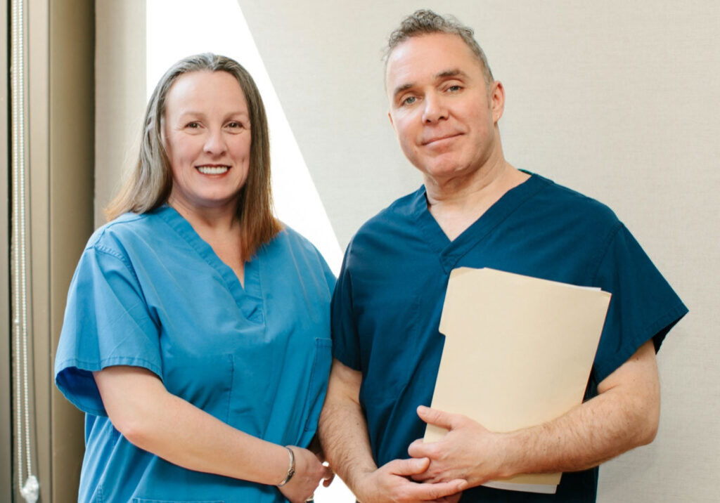 Two medical professionals in blue scrubs stand side by side, smiling. The person on the left has long brown hair, and the person on the right holds a file folder. They are indoors, in a well-lit room.