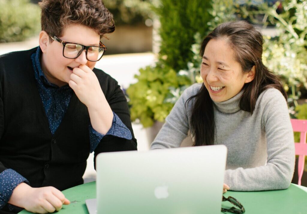 Two people sit at a green table outdoors, smiling and laughing while looking at a laptop. The person on the left wears glasses and a dark sweater; the person on the right wears a light gray turtleneck. Lush greenery is in the background.