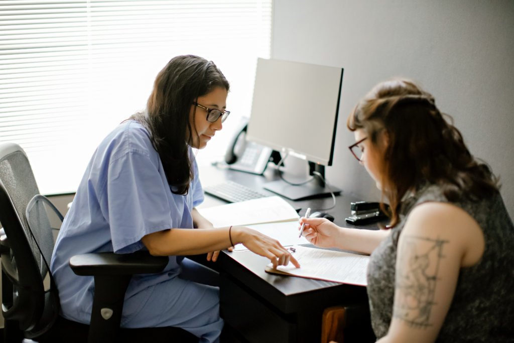 A healthcare worker in scrubs sits at a desk with a patient, reviewing documents together in a well-lit office with a computer and notepads.