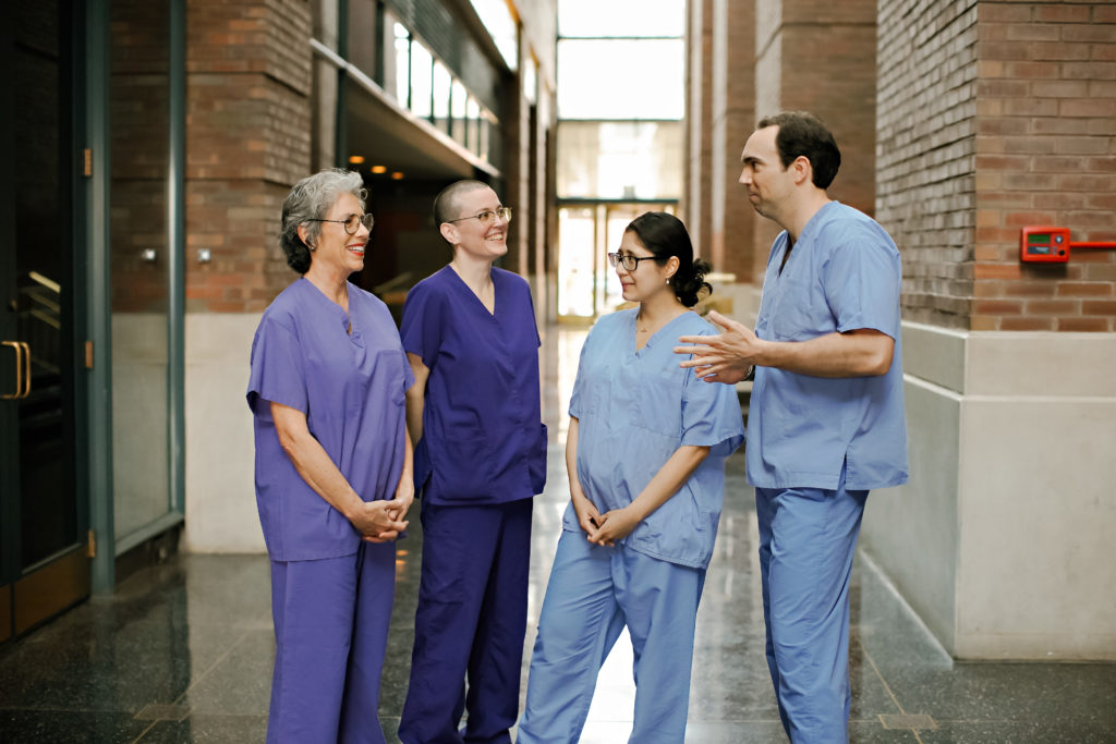 Four healthcare professionals in scrubs stand in a hallway with brick walls, smiling and talking together. Three are wearing purple scrubs and one is in blue scrubs. The setting appears to be a hospital or clinic.
