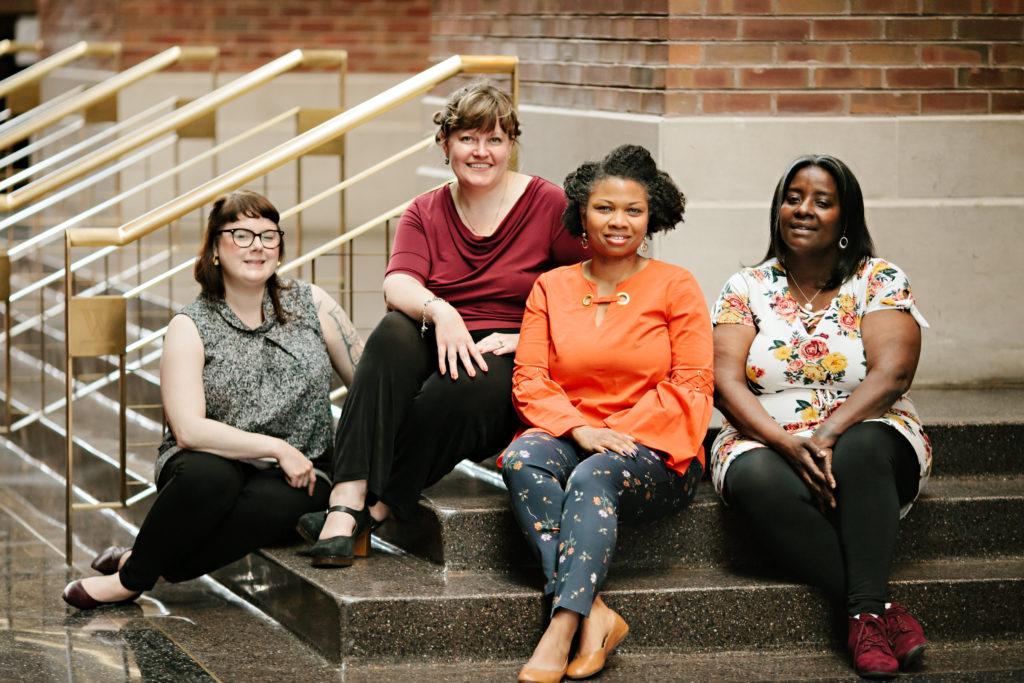 Four women sit together on indoor steps, smiling at the camera. The setting features brick walls and gold railings. The women wear a mix of casual and business-casual clothing in various colors and patterns.