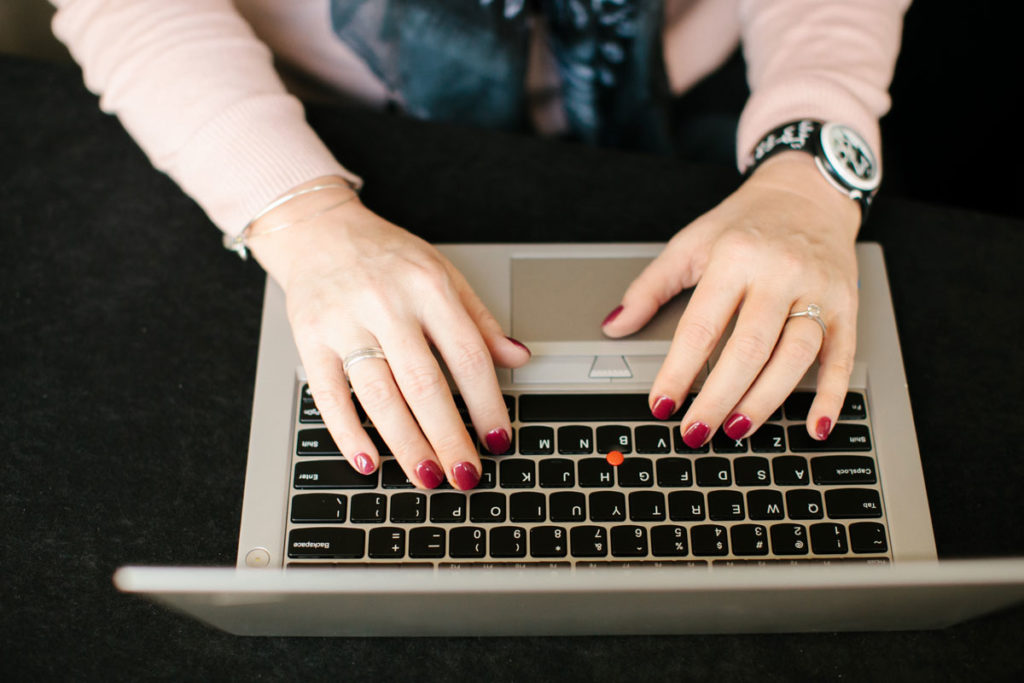 A person with painted nails types on a laptop keyboard. The individual is wearing a watch and bracelets, and the laptop is placed on a dark surface.